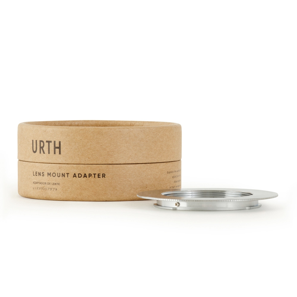 Urth M42 - Sony A (Minolta AF) lens adapter in a brown cylindrical box sits next to a silver metal lens mount adapter on a white background.