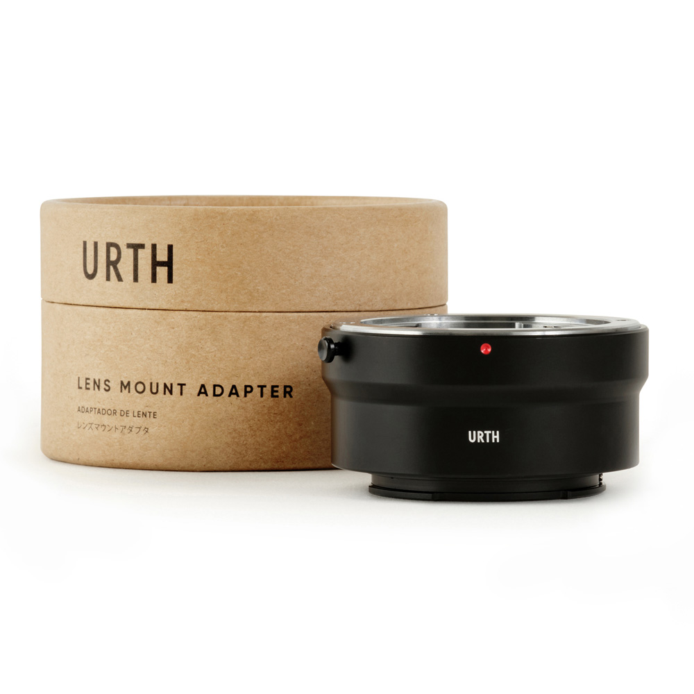 Musta Urth Contax/Yashica (C/Y) - Sony E objektiiviadapteri istuu sen pyöreän, ruskean pahvilaatikon vieressä, jossa on merkintä URTH Lens Mount Adapter, ja molemmat on sijoitettu tavalliselle valkoiselle taustalle.