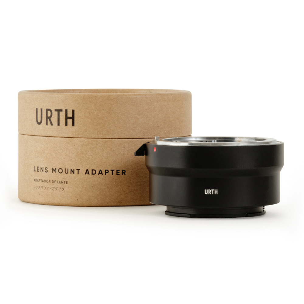 Musta Urth Nikon F - Sony E objektiiviadapteri näkyy lieriömäisessä laatikossa, jossa lukee "URTH Lens Mount Adapter", molemmat valkoista taustaa vasten.