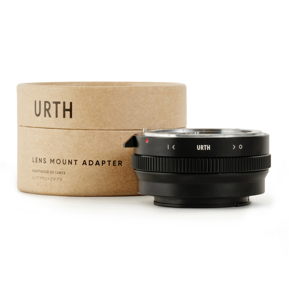 Musta Urth Nikon F (G-tyyppi) - Sony E objektiiviadapteri on sijoitettu sylinterimäisen pahvilaatikon eteen, jossa on merkintä URTH-objektiiviasennussovitin valkoisella pohjalla.