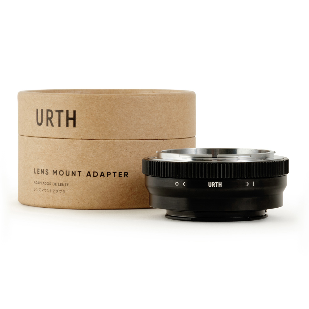 Ruskea lieriömäinen laatikko, jossa lukee "Urth Canon FD - Sony E objektiiviadapteri", on mustan metallisen, Urth-brändillä varustetun objektiiviasennussovittimen vieressä, molemmat valkoista taustaa vasten.
