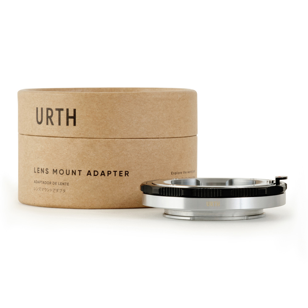 Pyöreä laatikko, jossa lukee "Urth Leica M - Sony E objektiiviadapteri (Extendable)", on metallisen objektiiviasennussovittimen vieressä, ja molemmat on sijoitettu valkoiselle taustalle.