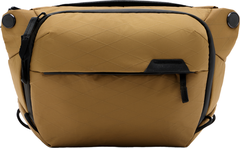 Peak Design Everyday Sling v2 3L - Coyote on ruskehtava, säänkestävä laukku, jossa on mustat korostukset, vetoketjullinen etutasku ja timanttikuvioinen kangas, joka näkyy yksiväristä taustaa vasten.