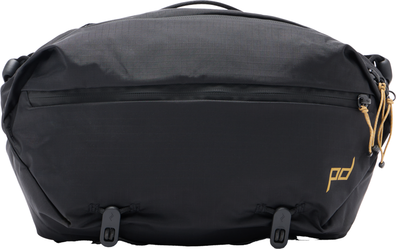 Peak Design Outdoor Sling 7L - musta -mallistossa on vetoketju edessä, kultainen logokirjoitus oikeassa alakulmassa, ruskehtavat vetoketjunvetimet oikealla puolella ja kaksi säädettävää hihnaa alareunassa.