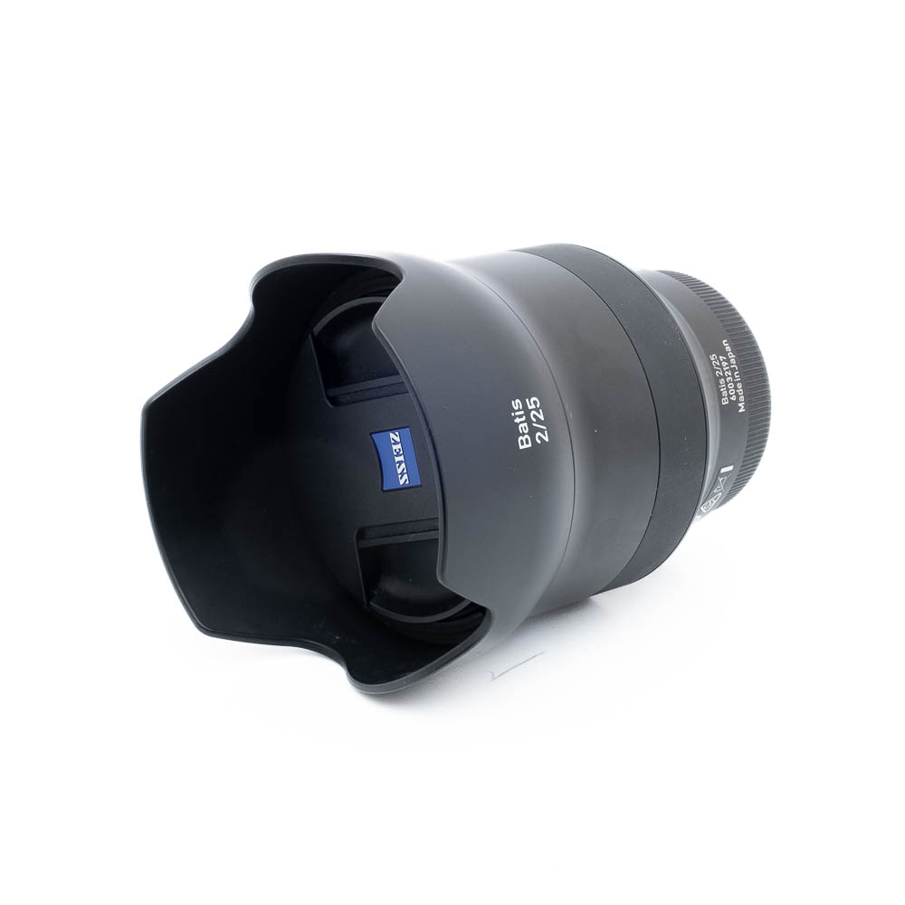 Mustan värinen Zeiss Batis 25mm f/2 Distagon Sony - Käytetty -objektiivi, jossa on terälehden muotoinen vastavalosuoja ja näkyvä sininen Zeiss-logo tynnyrissä, on kuvassa kyljellään valkoista taustaa vasten.