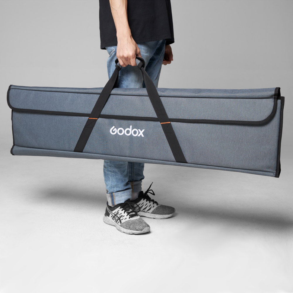 Farkkuihin ja lenkkareihin pukeutunut henkilö pitää kädessään suurta harmaata Godox Scrim Fast Flag Kit 60x90cm -kantolaukkua, jossa on mustat kahvat, harmaata taustaa vasten.