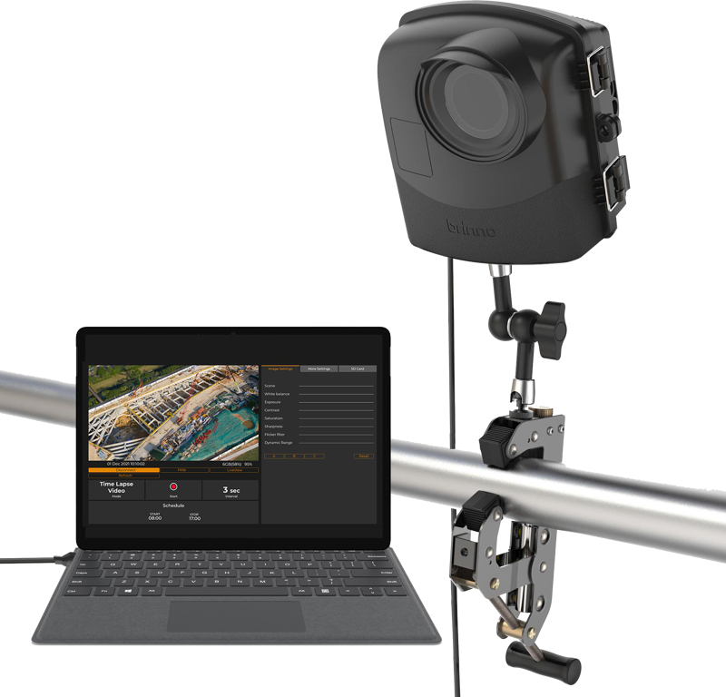 Mustan värinen Brinno BCC2000Plus Construction Camera Bundle on asennettu metallitolppaan ja liitetty kannettavaan tietokoneeseen, jossa näkyy editointiohjelmisto ja ilmakuva venesatamasta.