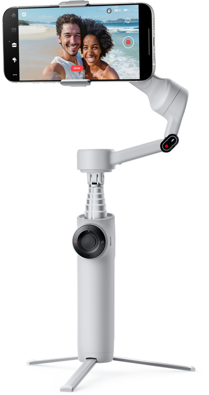 INSTA360 FLOW 2 STANDARD BUNDLE STONE GRAY -kardaani, jossa on kolmijalka, pitää älypuhelinta, jossa näkyy hymyilevä pariskunta ottamassa selfietä rannalla. Kivenharmaassa gimbalissa on säätöpyörä ja pidennetty kahva.